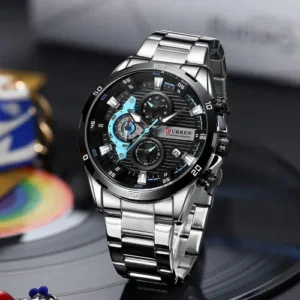 Reloj Curren 8402 Plateado, cronógrafo con caja plateada y esfera negra. Presenta múltiples sub-diales con detalles en turquesa/azul claro y plateado. La esfera muestra el logo Curren en rojo y tiene un indicador de fecha. El reloj cuenta con una correa metálica de eslabones en acero inoxidable plateado. Se muestra en ángulo sobre una superficie oscura, resaltando su diseño deportivo y elegante con bisel numerado
